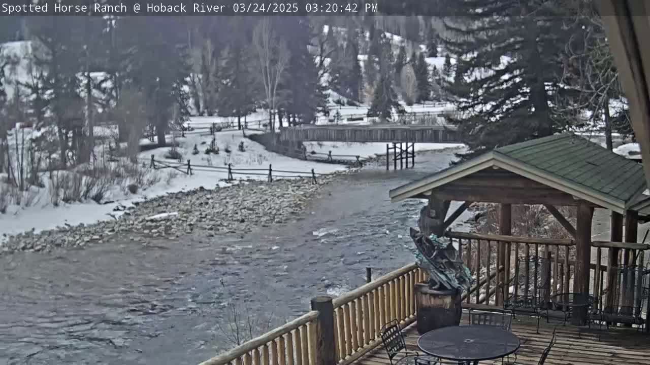 A snow-dusted river flows past a wooden deck with a gazebo and outdoor furniture, under a partly cloudy sky.