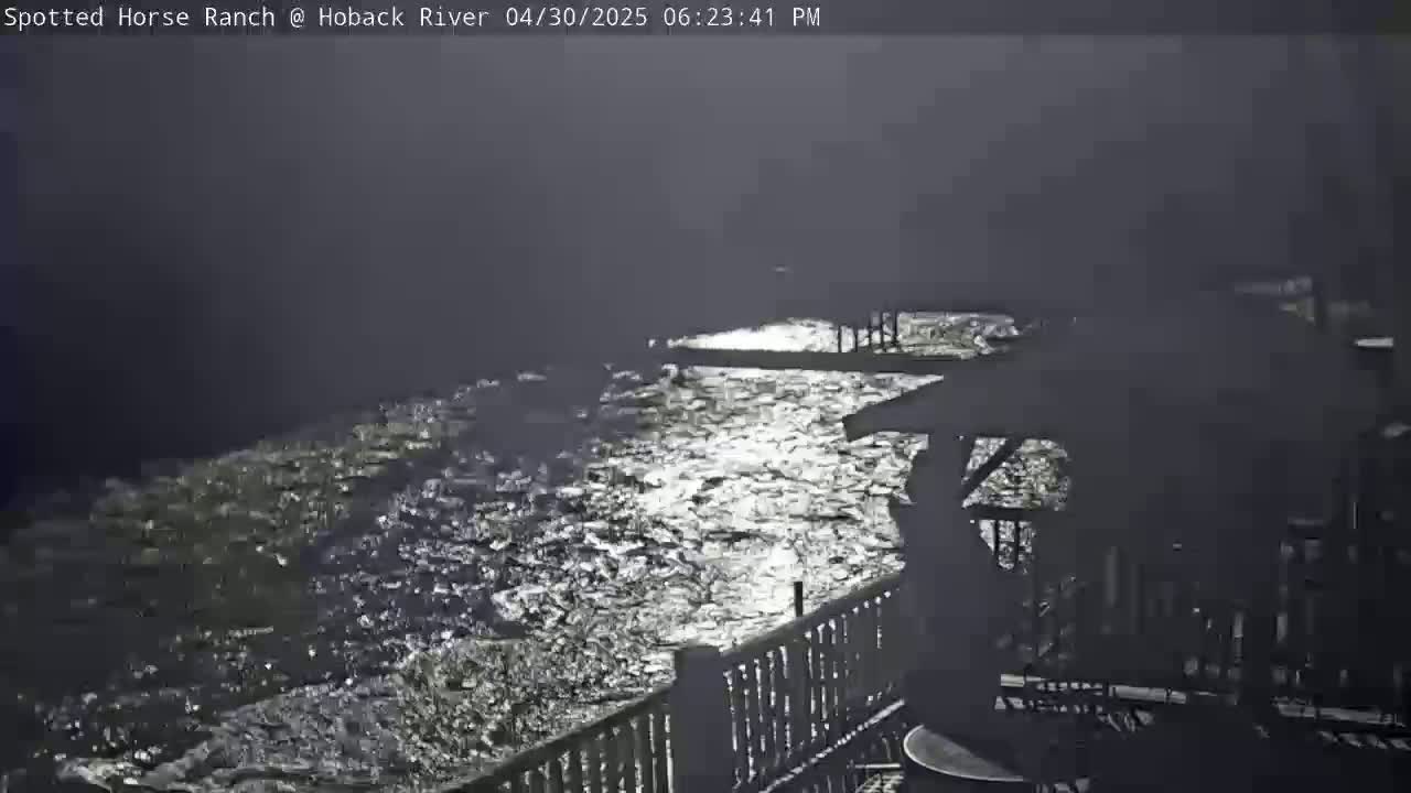 A nighttime view from a deck shows a swiftly flowing river reflecting moonlight under a dark, cloudy sky.