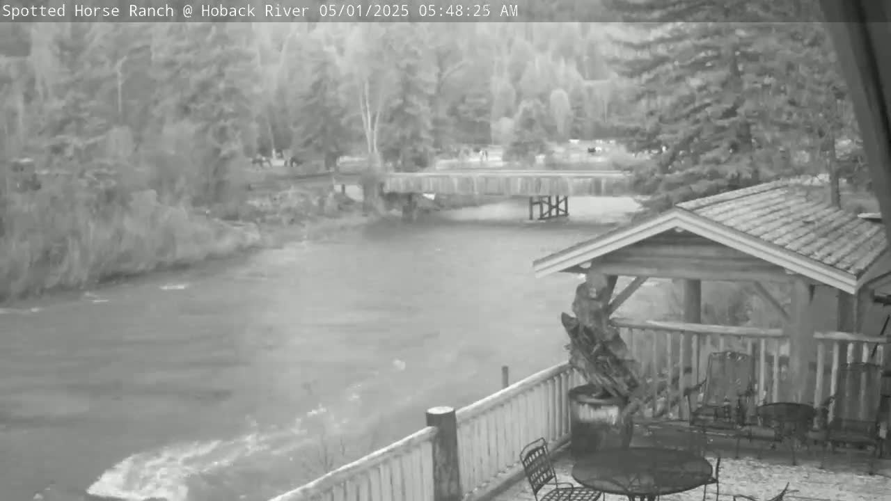 A river flows under a bridge near a wooden deck with patio furniture, all surrounded by trees in the early morning.