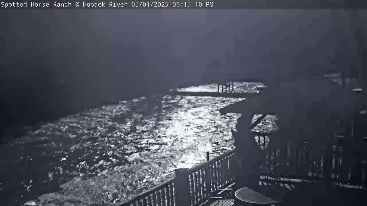 A nighttime view shows a dark, swiftly flowing river reflecting light, seen from a deck with a railing.