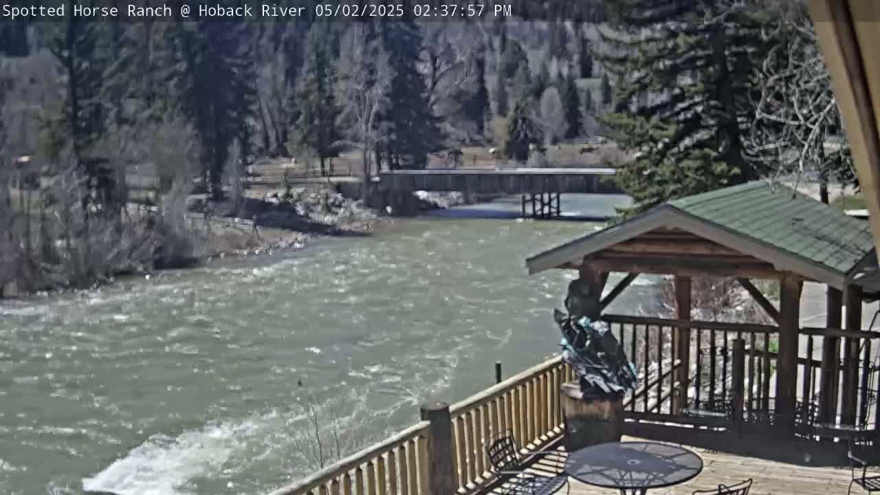 A deck overlooking a swiftly flowing river, with a wooden gazebo and a statue, on a sunny day.