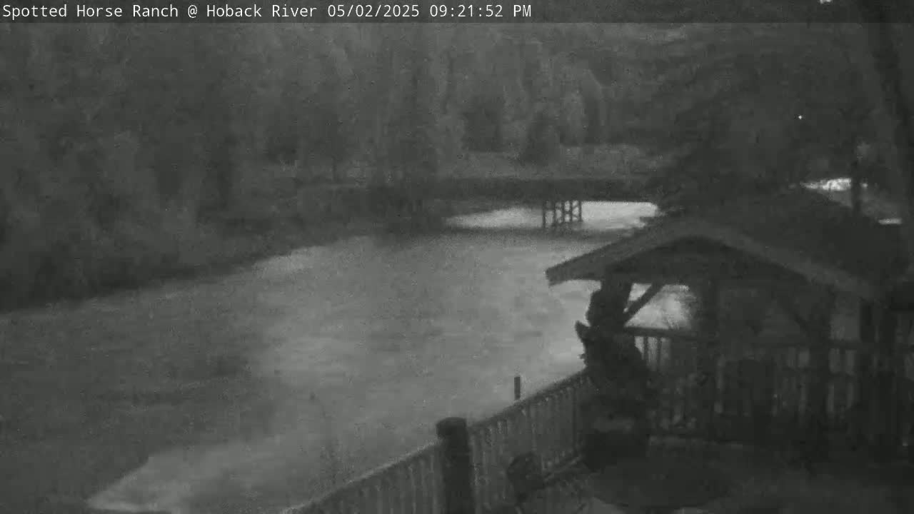 A nighttime, low-light image shows a turbulent river flowing beneath a covered structure with a railing, partially obscuring the view of the water.