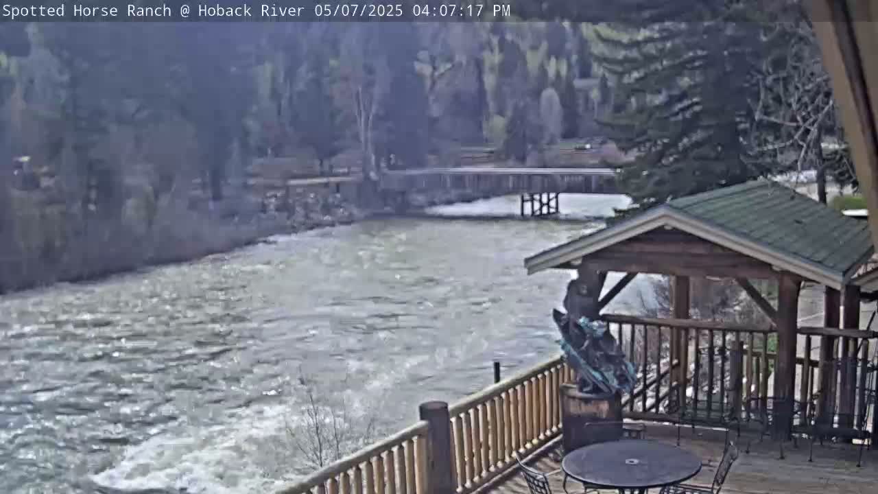 A rushing river flows beneath a wooden deck with a gazebo, overlooking a bridge and wooded area.