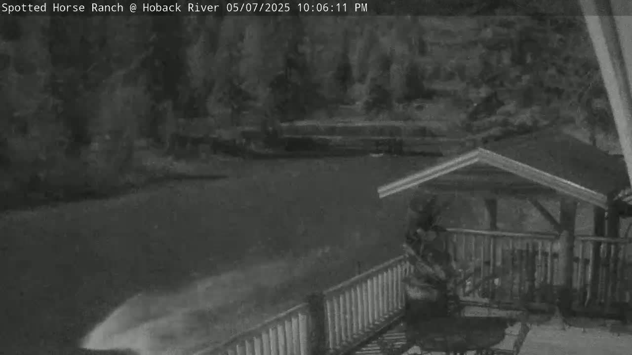 A nighttime, low-light view shows a partially snow-covered deck with a railing, a small structure, and a dark, indistinct area beyond.