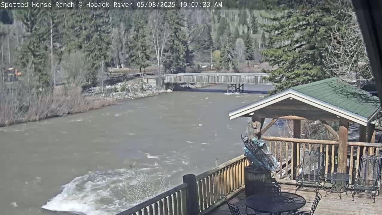 A deck with a gazebo overlooks a swiftly flowing river and a wooden bridge under a mostly sunny sky.