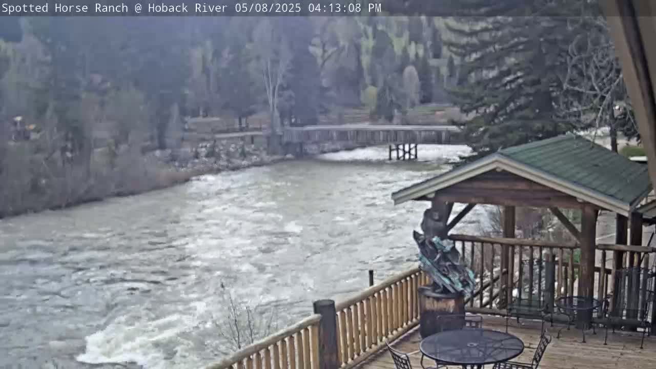 A fast-flowing river is seen from a wooden deck with a gazebo and outdoor furniture, under an overcast sky.
