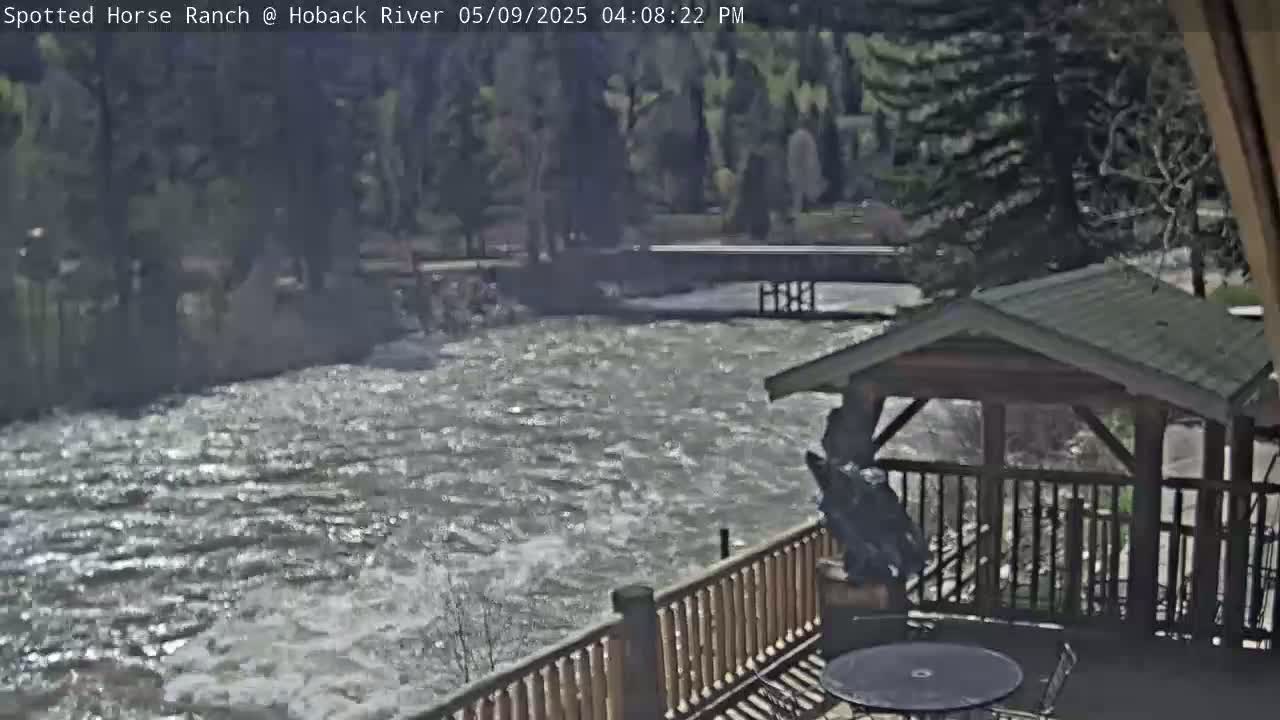 A fast-flowing river is seen from a deck with a wooden gazebo and railing, under a sunny sky.