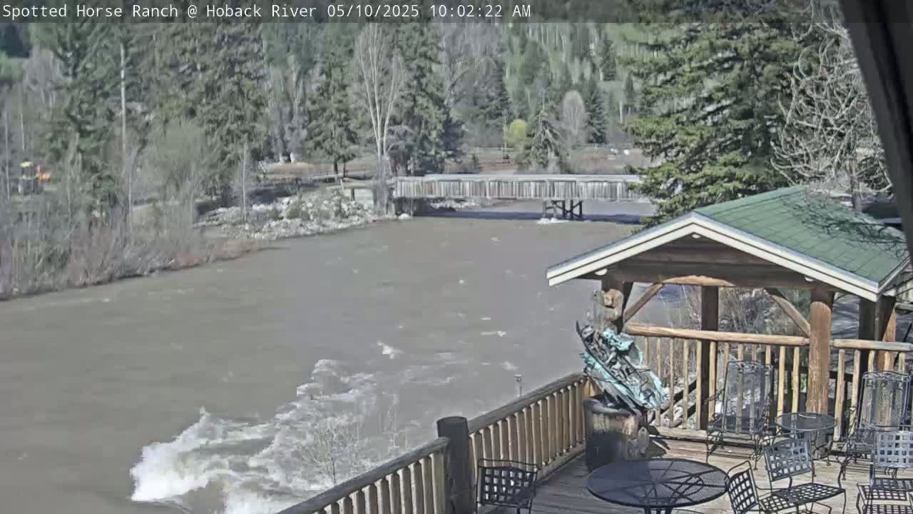 A fast-flowing river is viewed from a wooden deck with a gazebo and outdoor furniture under a partly sunny sky.