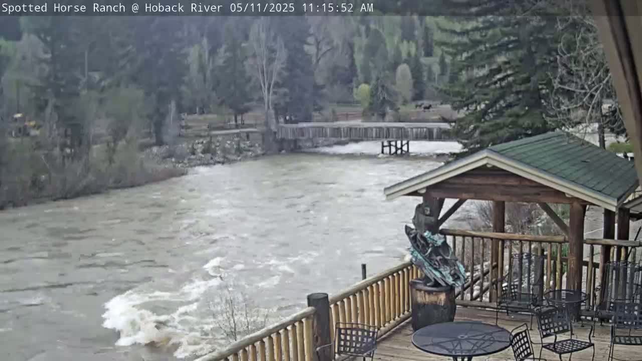 A rushing river flows beneath a wooden deck with outdoor furniture, next to a bridge and wooded area.