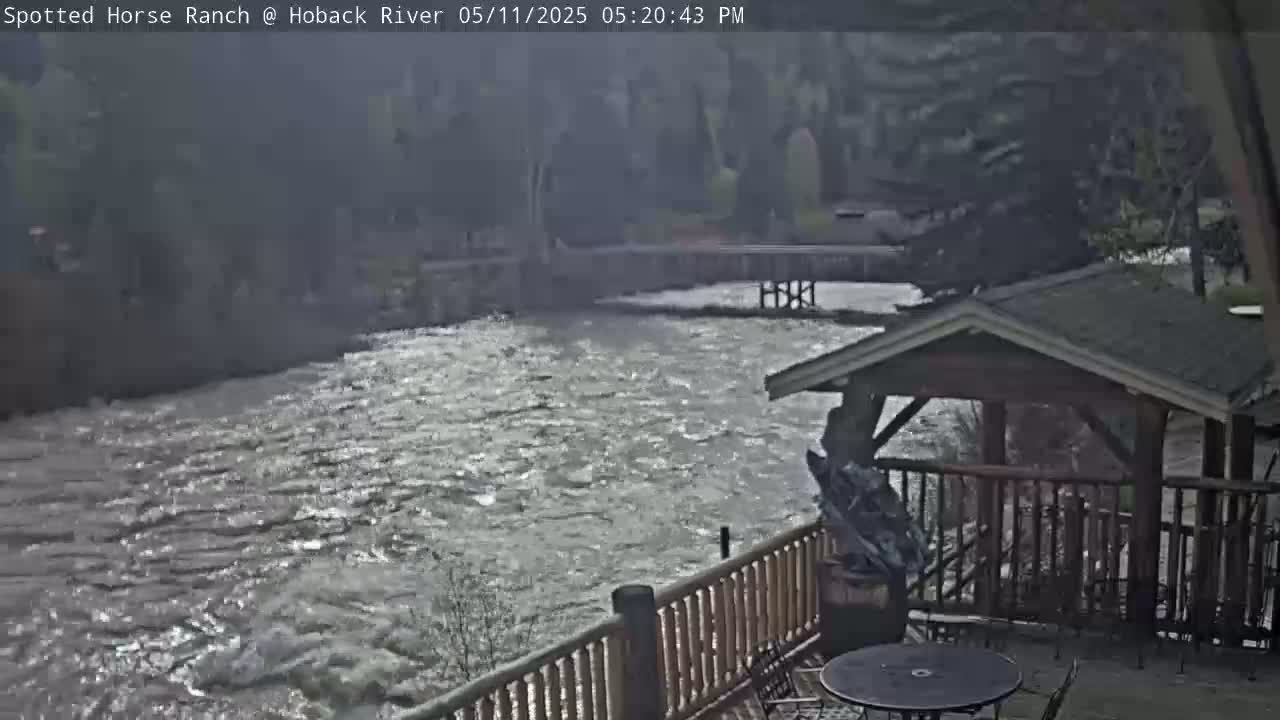 A fast-flowing river is viewed from a wooden deck with a gazebo, under a cloudy sky.