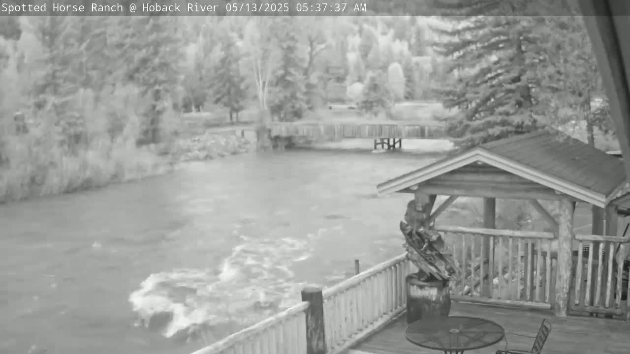 A fast-moving river flows beneath a wooden deck and gazebo, with trees lining the riverbank in the background.