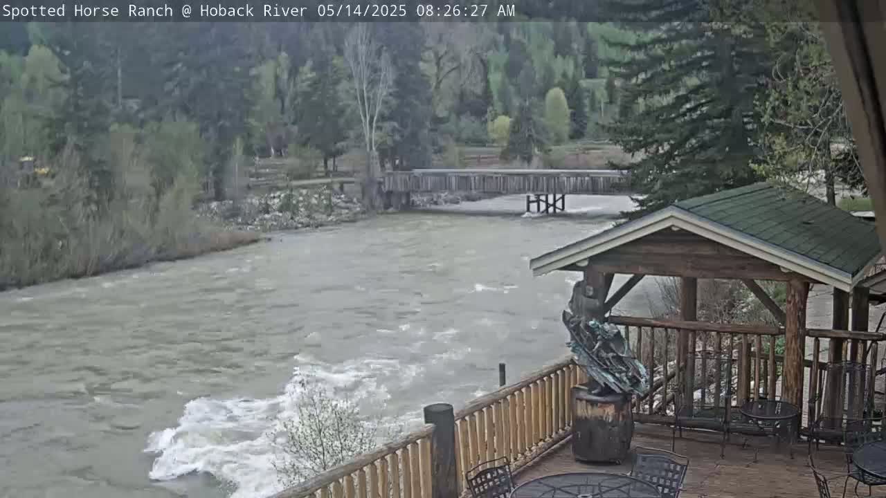 A fast-flowing, muddy river rushes beneath a wooden deck and gazebo, with a wooden bridge visible in the background under an overcast sky.