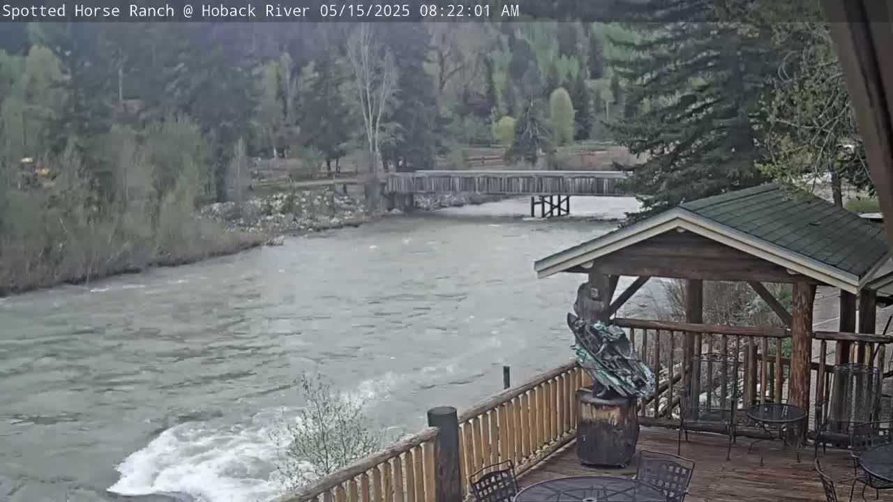 A fast-moving river flows under a wooden bridge, visible from a wooden deck with outdoor furniture and a sculpture; the weather appears overcast.
