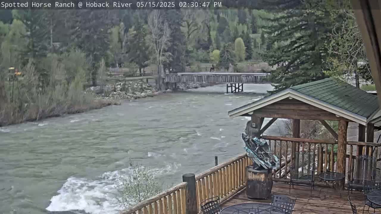 A fast-flowing river is seen from a wooden deck with a gazebo,  surrounded by green trees and a wooden bridge in the background on an overcast day.