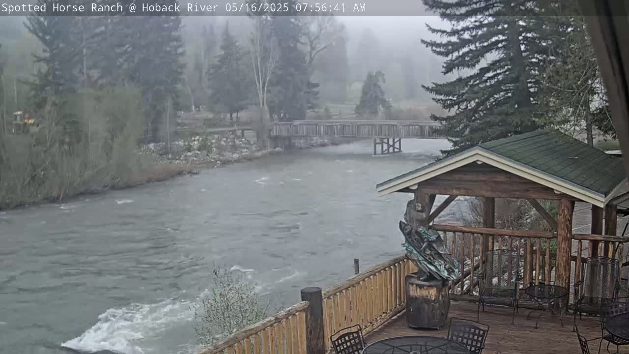 A fast-moving river flows under a wooden bridge, viewed from a deck with a gazebo and outdoor furniture, in foggy conditions.