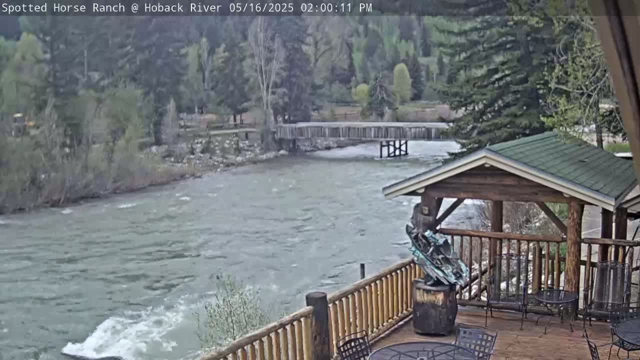 A deck overlooking a rushing river with a wooden bridge in the background, under partly cloudy skies.