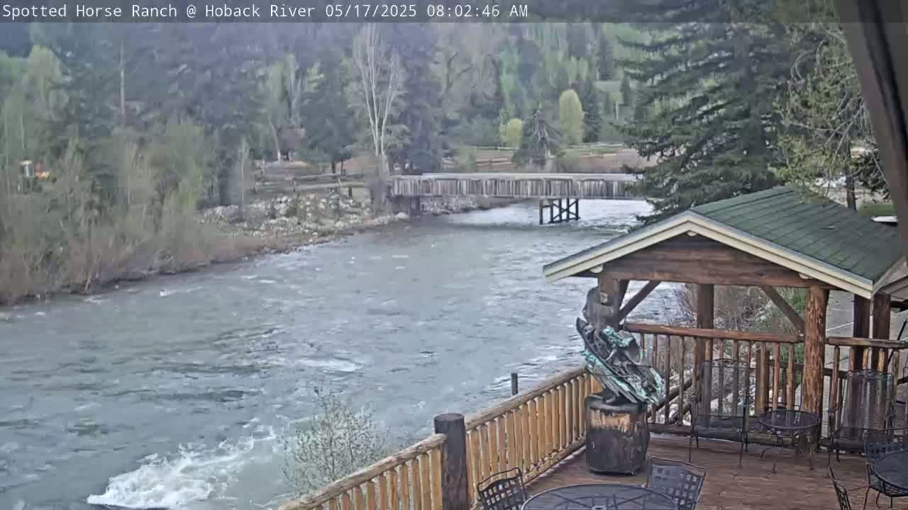 A wooden deck with a gazebo overlooks a flowing river and wooden bridge, with trees and hills visible in the background under overcast skies.