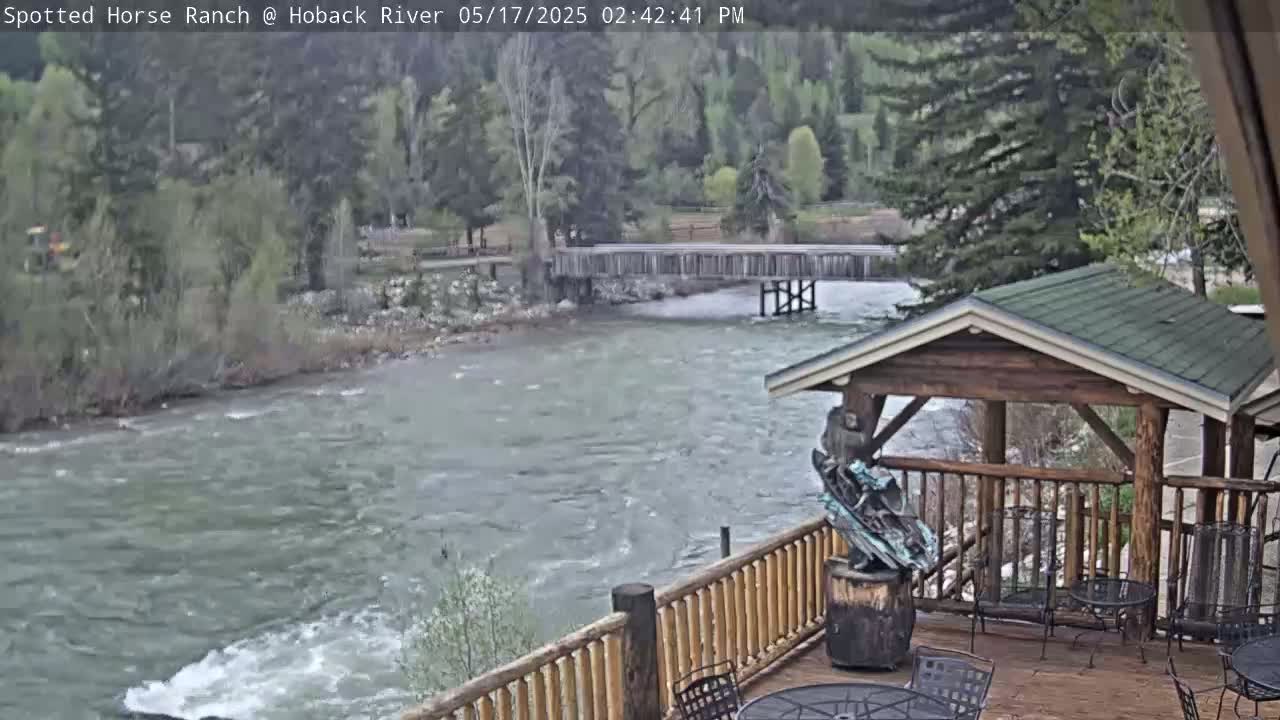 A rushing river flows under a wooden bridge, visible from a deck with wooden railings and outdoor seating, partially shaded by a gazebo-like structure.