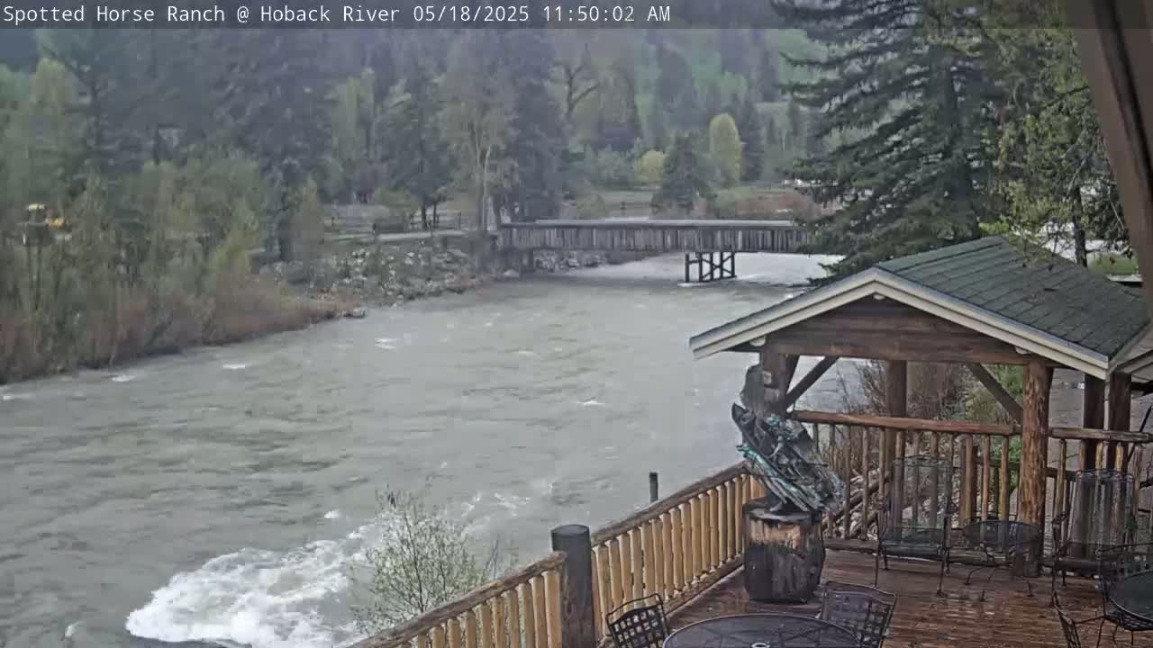 A turbulent river flows under a wooden bridge, viewed from a deck with a wooden gazebo and outdoor seating in rainy weather.