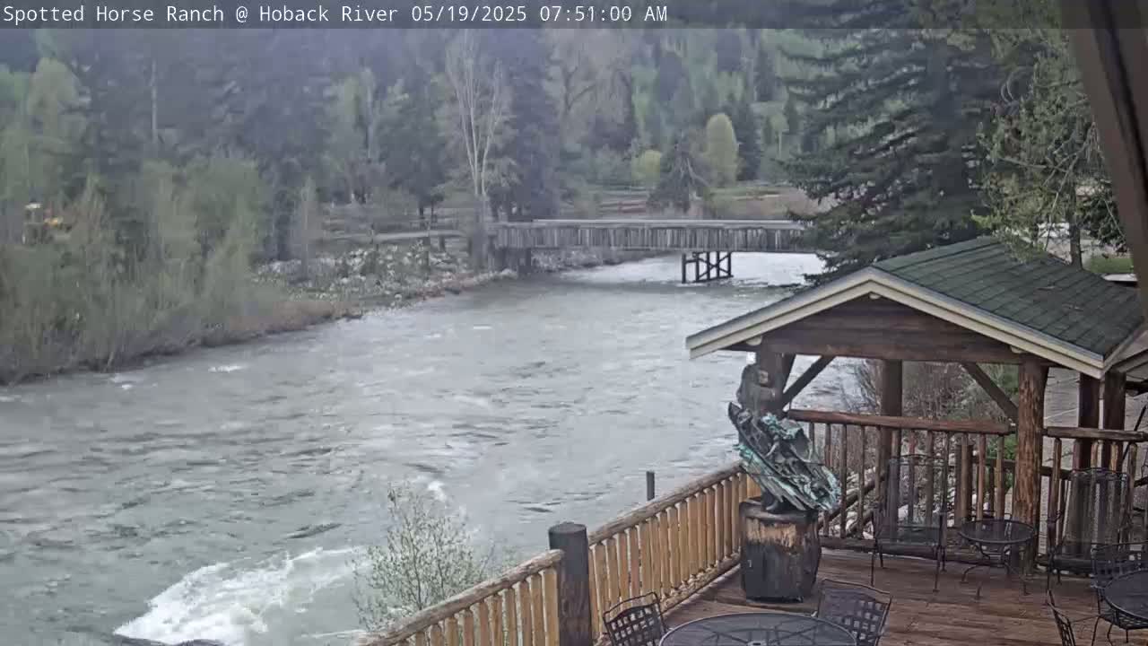 A deck with wooden railings overlooks a flowing river and a wooden bridge under an overcast sky.