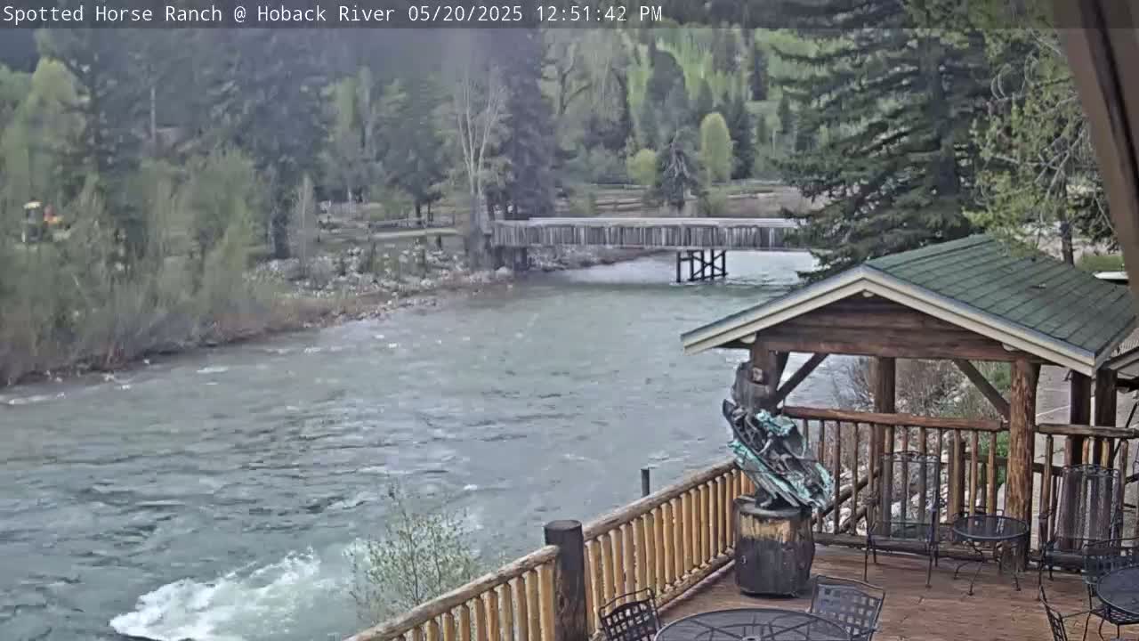 A partially overcast view of a flowing river with a wooden footbridge in the background, seen from a wooden deck with a gazebo and outdoor seating.