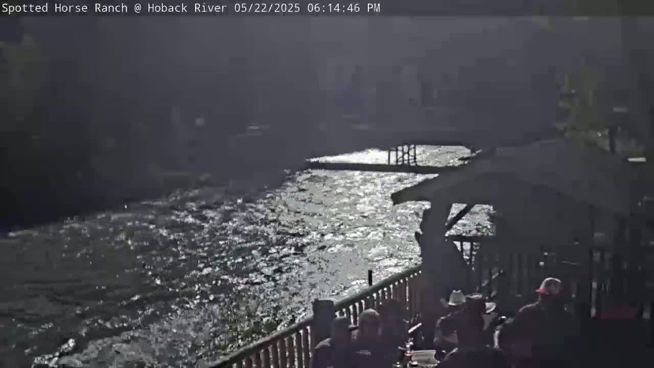A fast-moving river reflects sunlight, with several people sitting at an outdoor deck overlooking it.