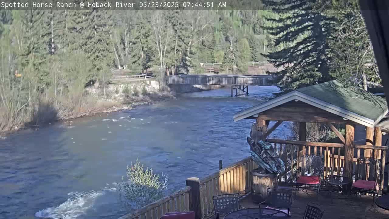 A flowing river, under a mostly sunny sky, is viewed from a wooden deck with a gazebo, overlooking a wooden bridge.