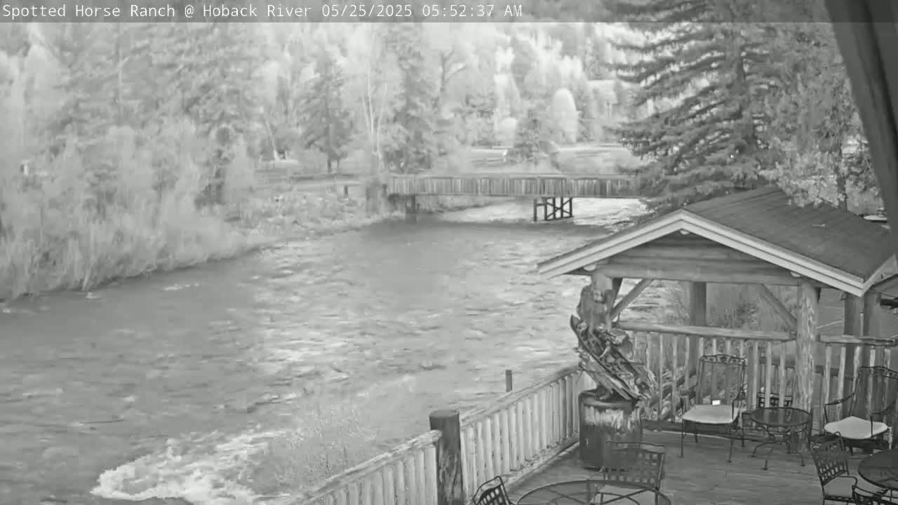 A fast-flowing river runs under a wooden bridge, viewed from a deck with a wooden gazebo and several empty chairs.