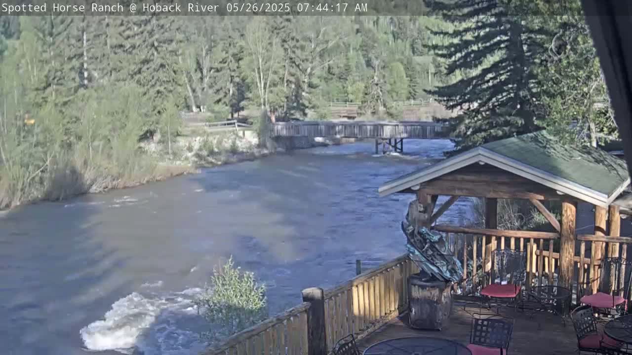 A fast-flowing river is seen from a wooden deck with a gazebo and outdoor seating, surrounded by trees under a partly sunny sky.