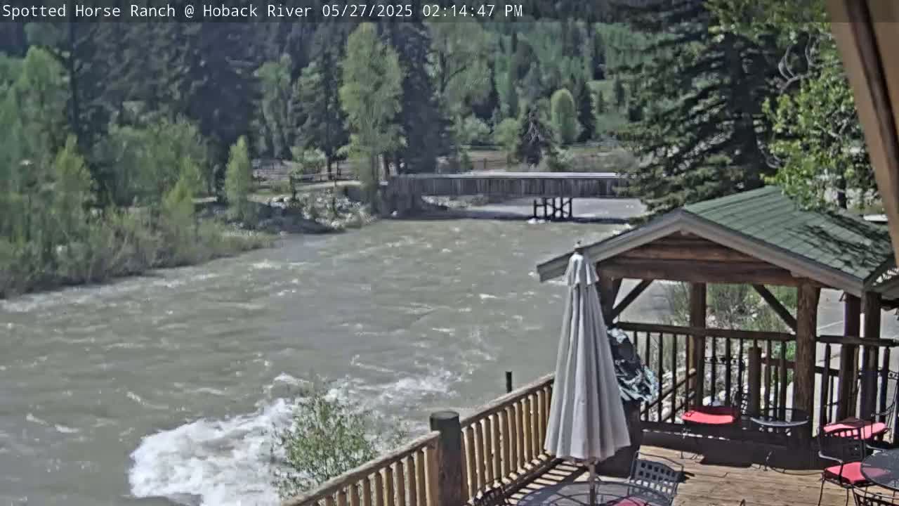 A swiftly flowing river is viewed from a wooden deck with a gazebo and patio furniture, under a sunny sky.