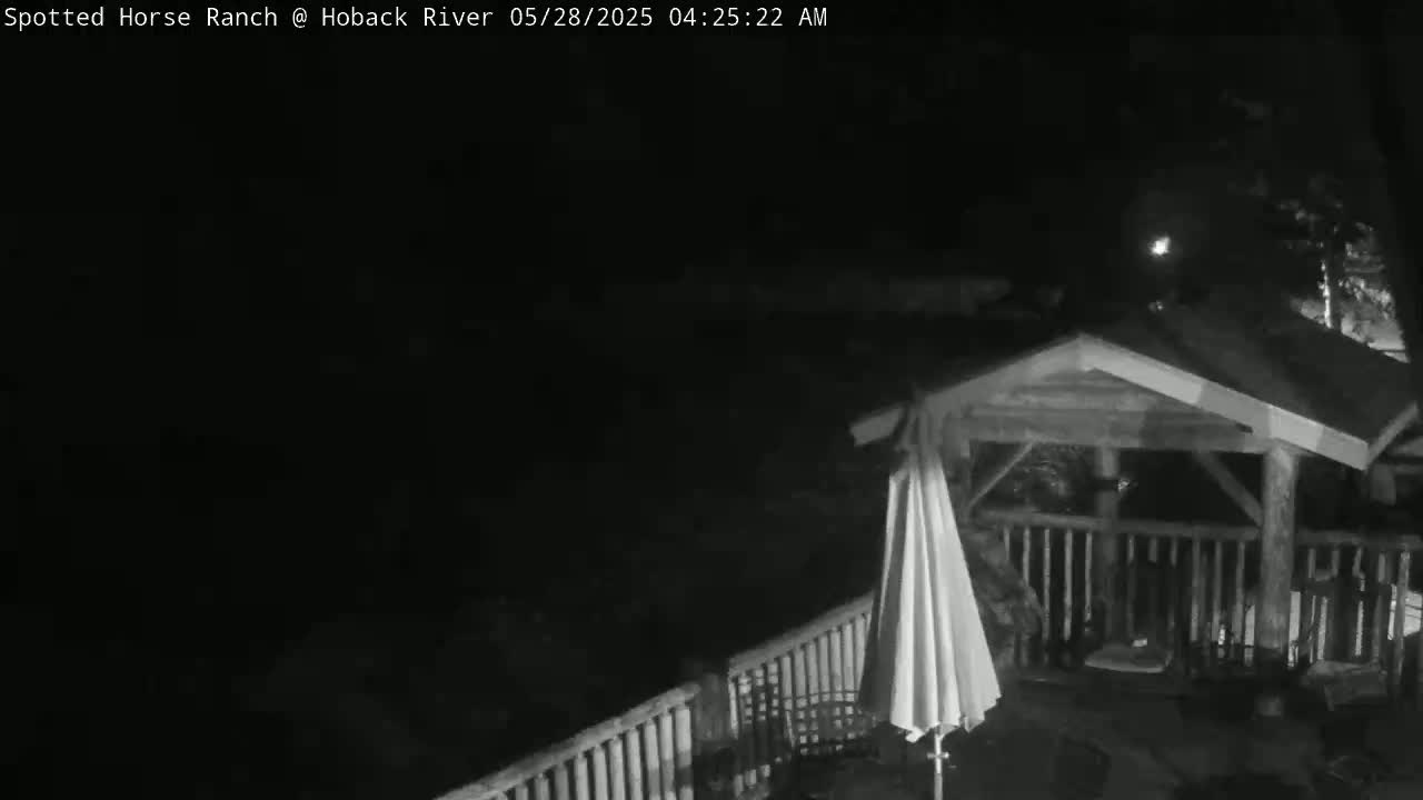 A nighttime, black-and-white image shows a wooden gazebo with a closed umbrella on a deck, partially illuminated by a light source in the distance.