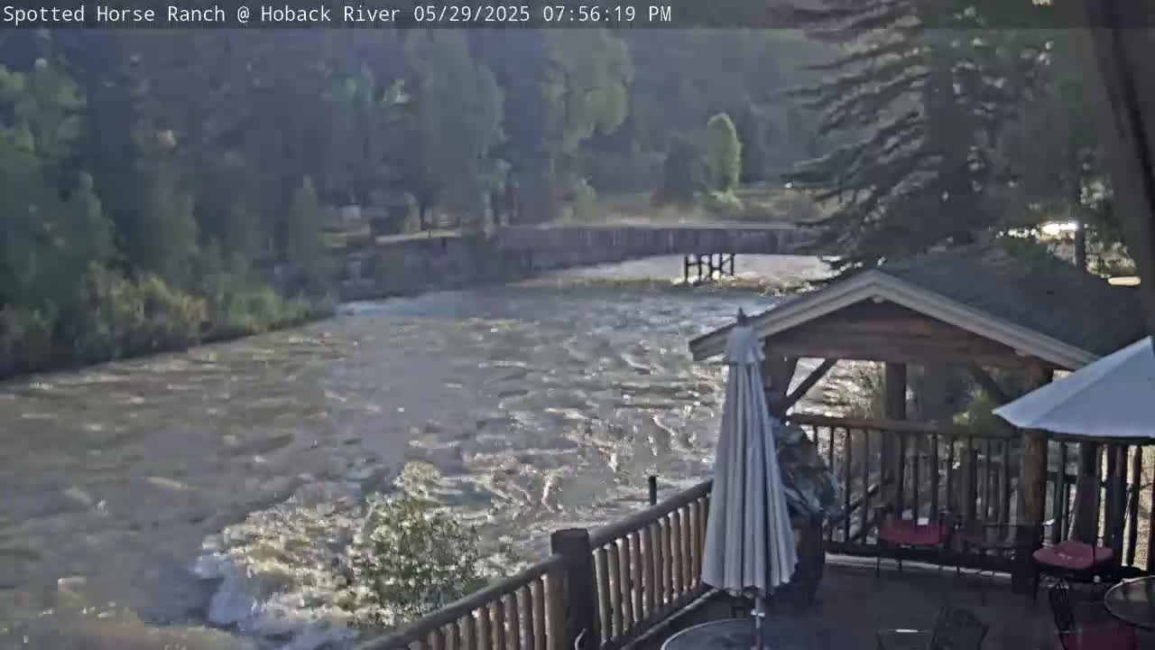 A fast-flowing river is visible from a wooden deck with a gazebo and patio umbrellas, under a partly cloudy sky.