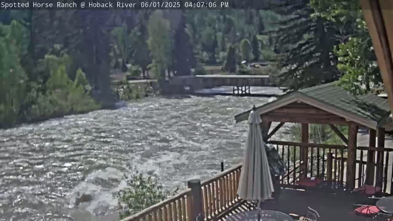 A swiftly flowing river is viewed from a wooden deck with a gazebo, under a partly sunny sky.