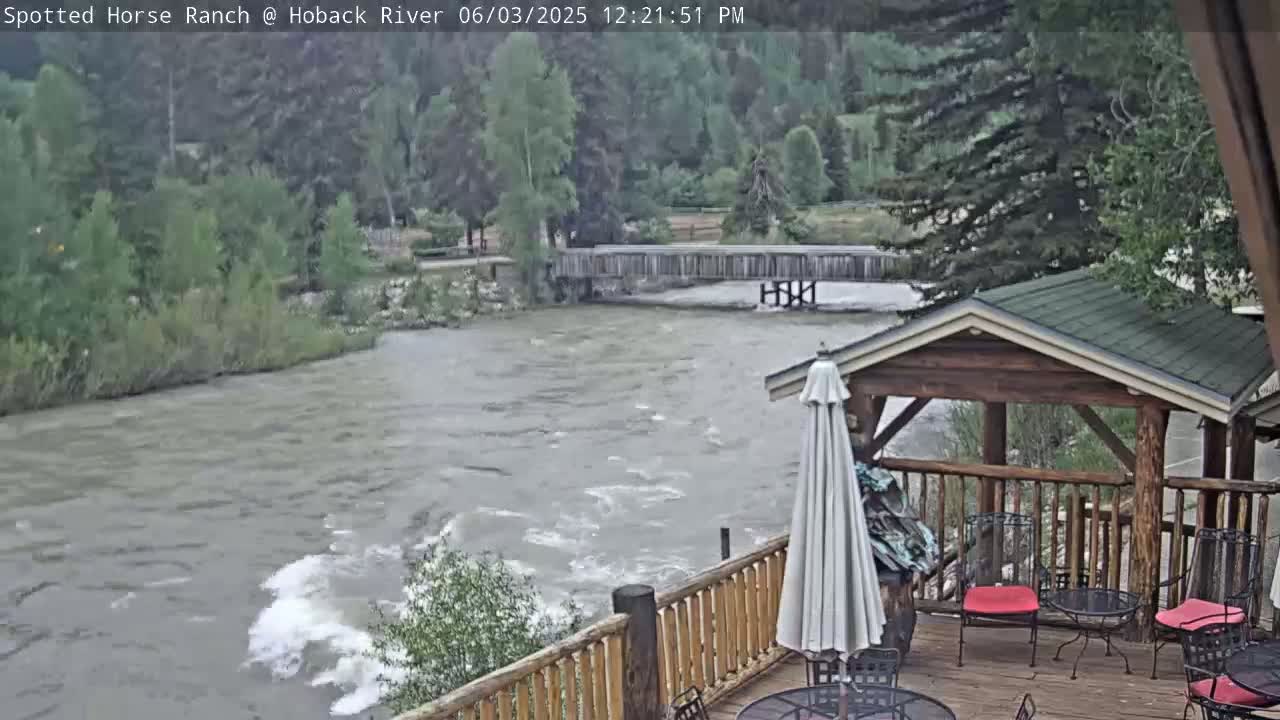 A swiftly flowing river is viewed from a wooden deck with a gazebo and outdoor seating, under overcast skies.