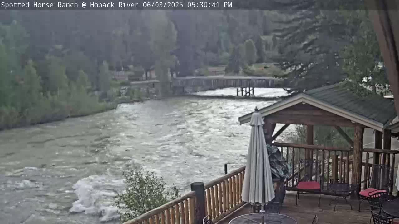 A rushing river flows beneath a wooden deck with a gazebo, under an overcast sky.