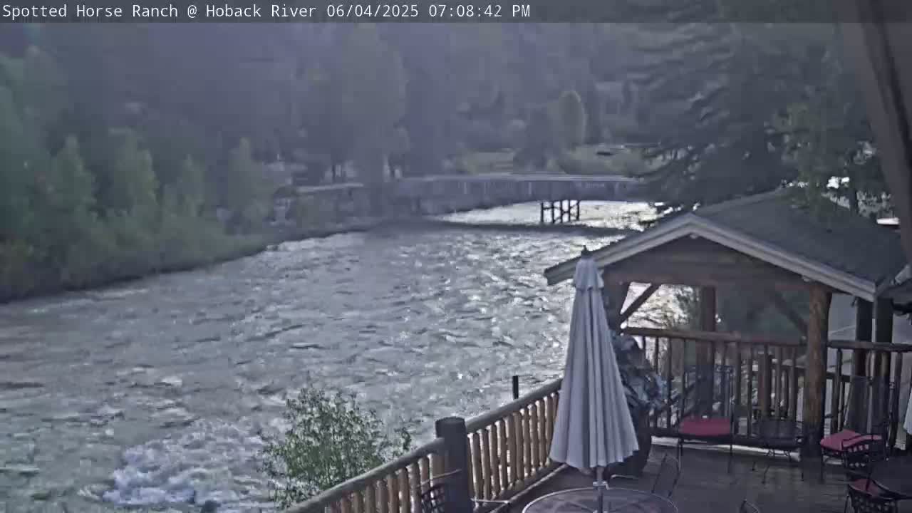 A deck with patio furniture overlooks a swiftly flowing river and a bridge, under a somewhat overcast sky.