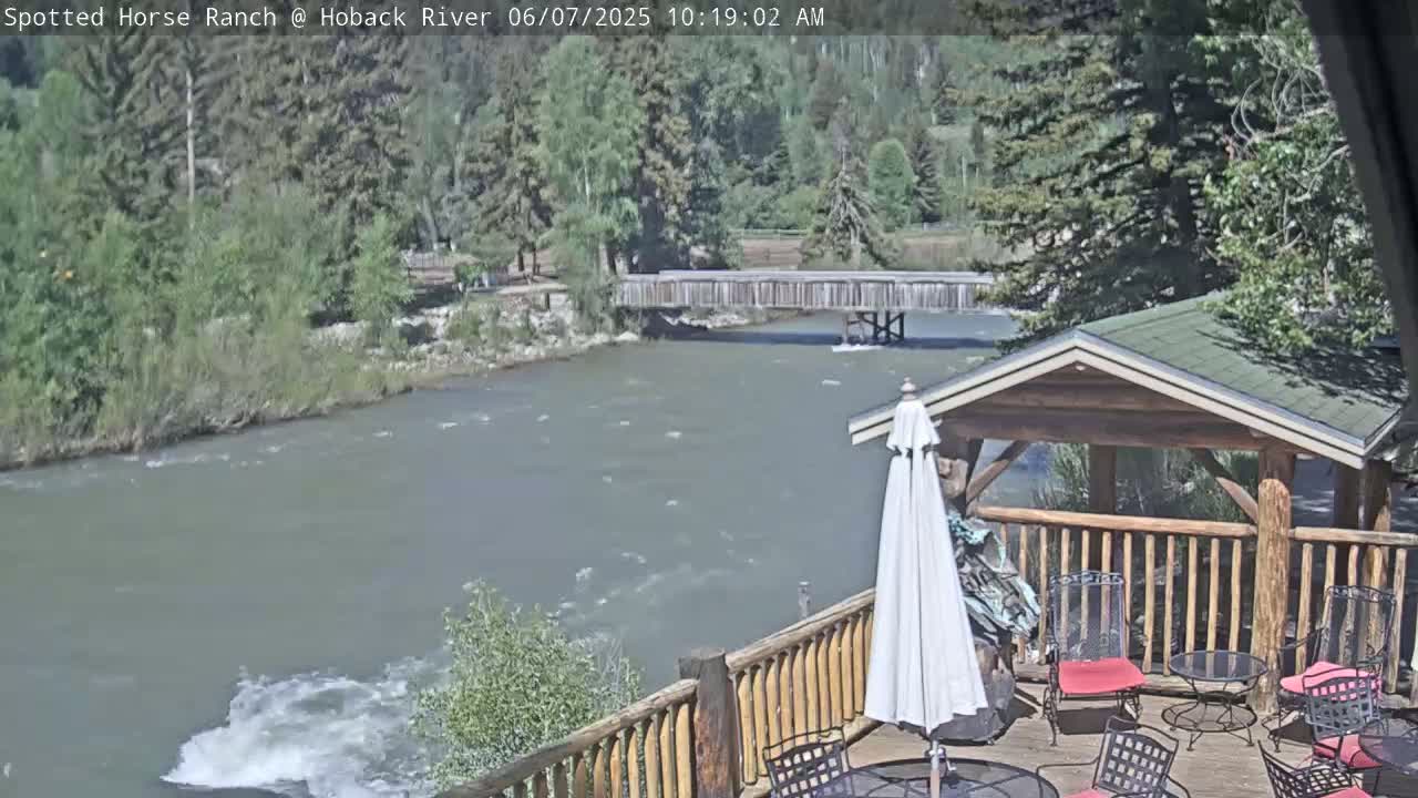 A wooden deck with patio furniture overlooks a flowing river and a wooden bridge, under a partly sunny sky.