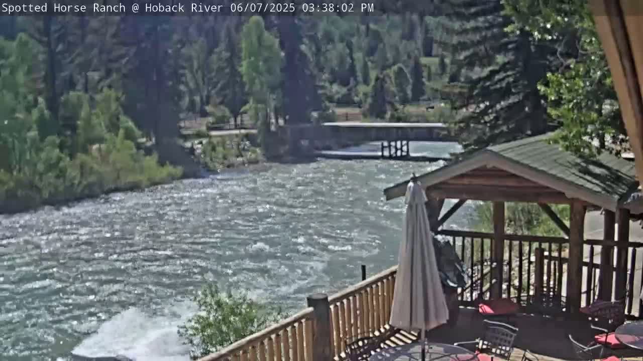 A wooden deck with a gazebo overlooks a swiftly flowing river on a sunny day, surrounded by green trees and vegetation.