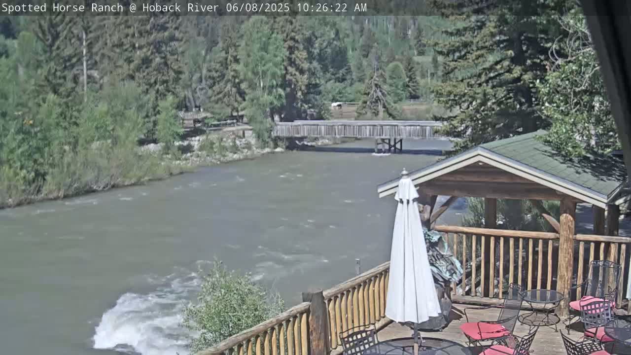 A wooden deck with patio furniture overlooks a swiftly flowing river and a wooden bridge under a mostly sunny sky.