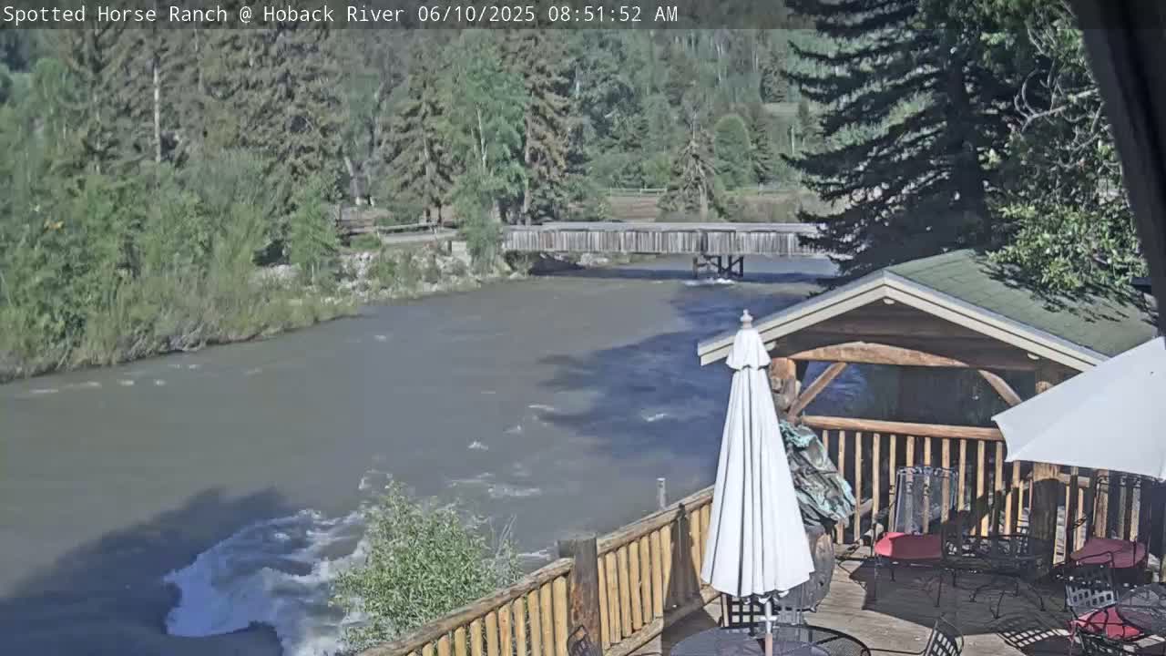 A partly sunny view of a river flowing under a wooden bridge, with a wooden deck and patio furniture in the foreground.
