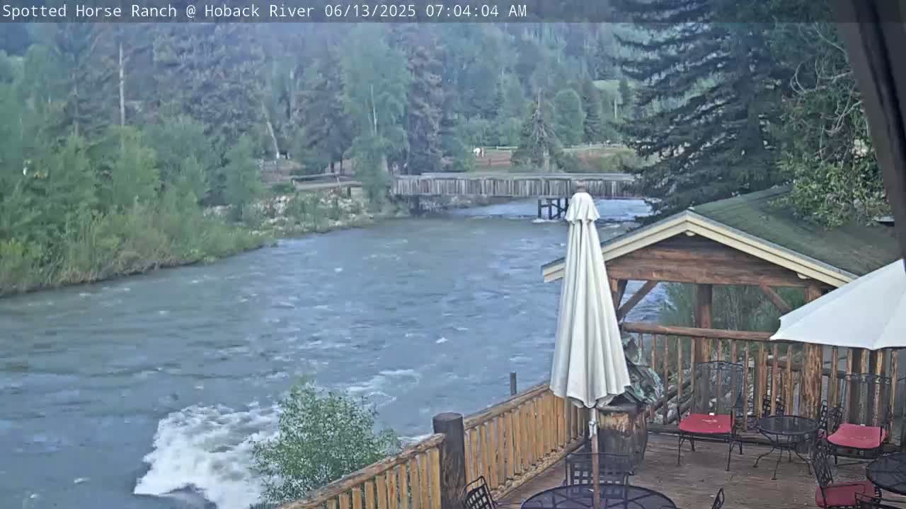 A deck with patio furniture overlooks a swiftly flowing river, bordered by green trees and a wooden footbridge under a mostly cloudy sky.