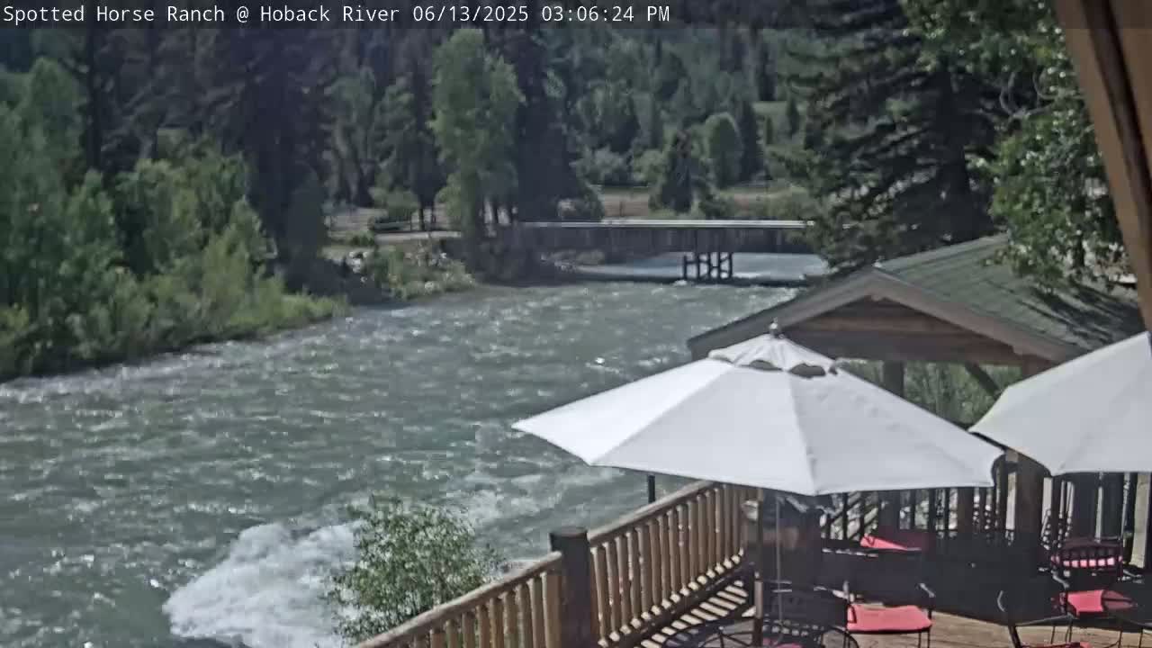 A deck with patio umbrellas overlooks a swiftly flowing river on a sunny day, with lush green trees lining the riverbank in the background.