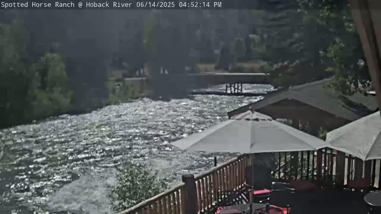 A deck with patio furniture and umbrellas overlooks a flowing river and a wooden footbridge, under a mostly sunny sky.