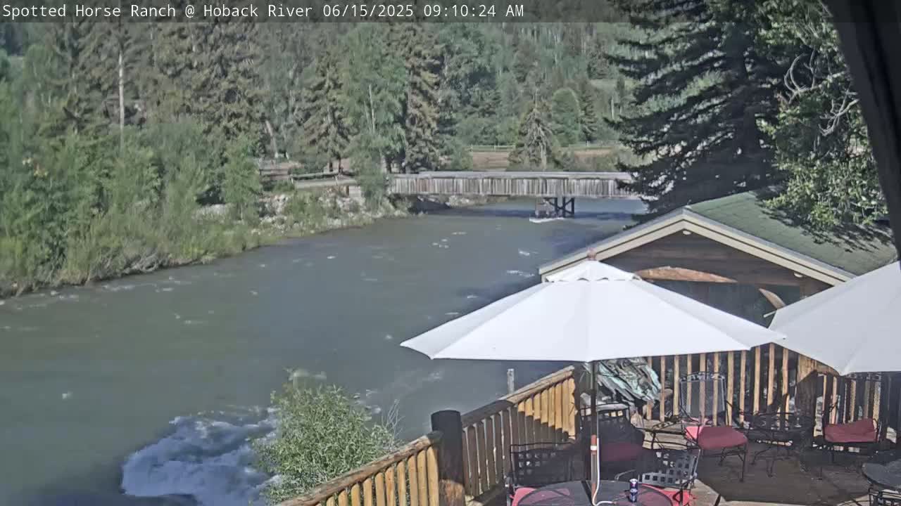 A deck with patio furniture and umbrellas overlooks a flowing river and wooden bridge under a sunny sky.