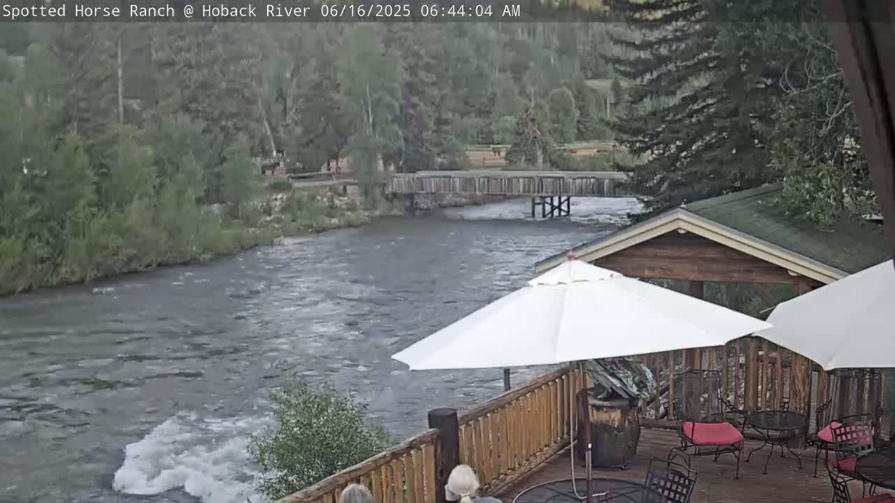 A deck with patio furniture overlooks a swiftly flowing river and wooden footbridge, all under an overcast sky.