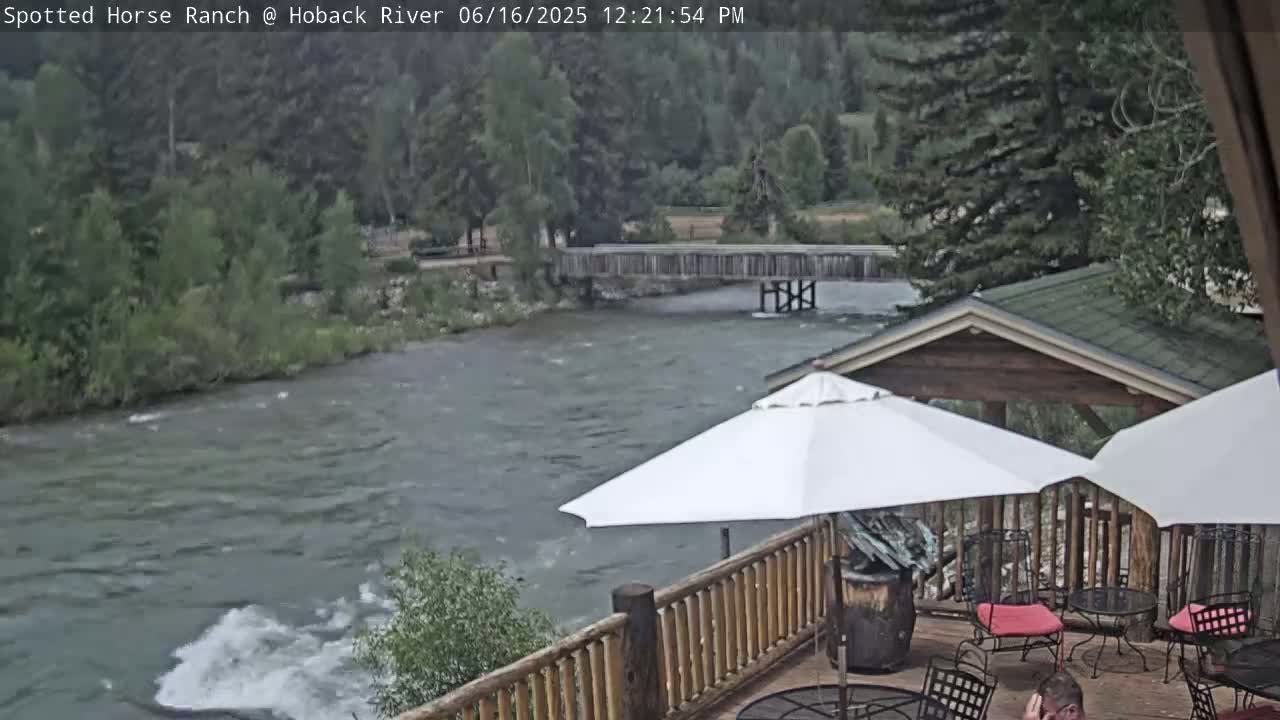 A turbulent river flows beneath a wooden footbridge, past a patio with umbrellas and outdoor seating under an overcast sky.