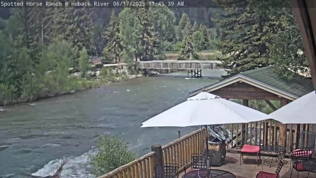 A deck with patio furniture and umbrellas overlooks a flowing river and a wooden footbridge under a partly cloudy sky.