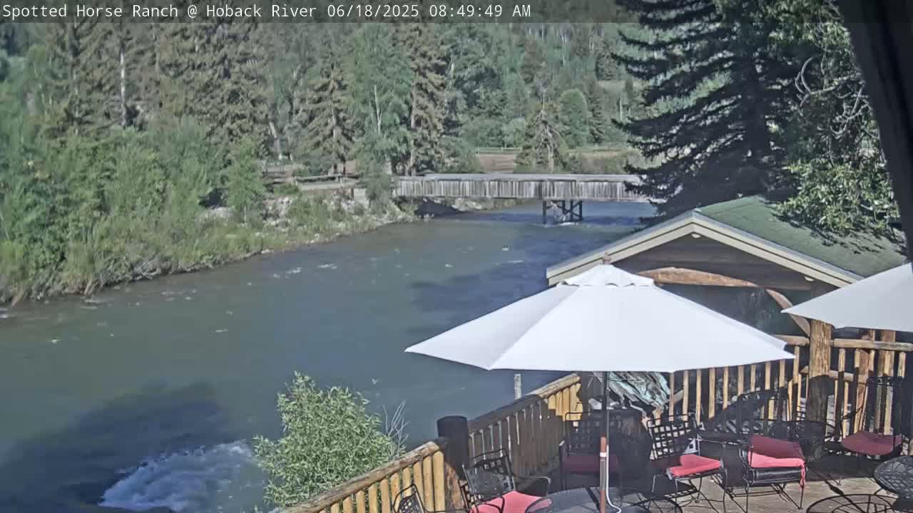 A patio with umbrellas overlooks a river on a sunny day, with a wooden bridge visible in the distance.