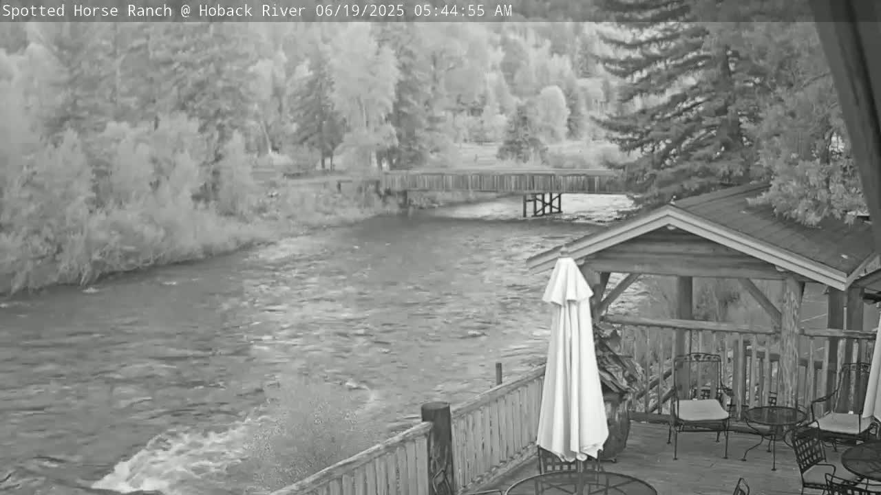 A fast-moving river flows under a wooden bridge, past a deck with patio furniture and a closed umbrella, surrounded by trees.