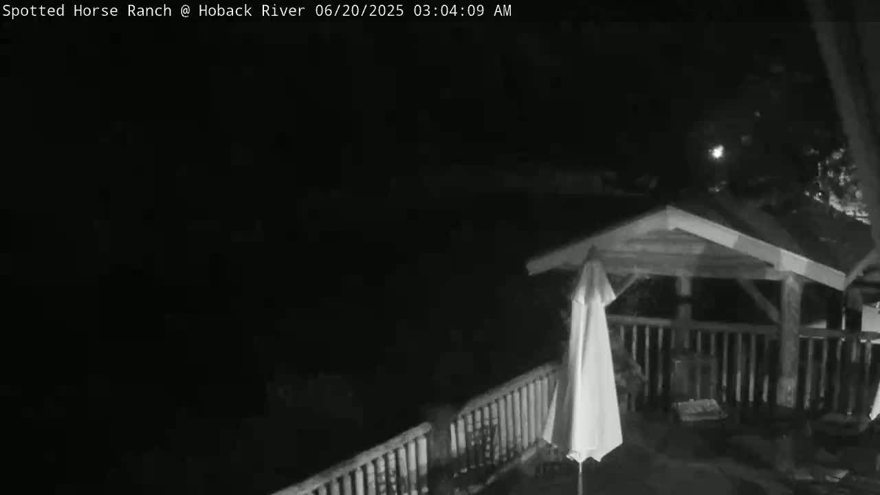 A nighttime, low-light image shows a wooden gazebo with a railing and a closed umbrella on a deck.